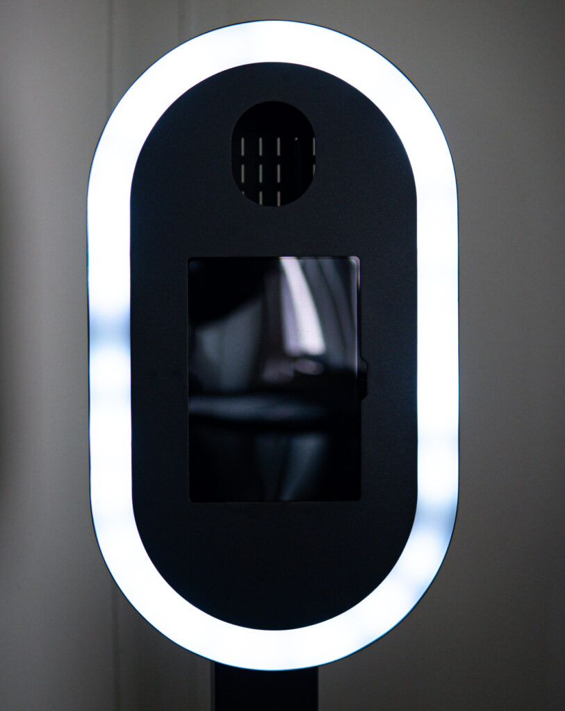 Modern photo booth with an illuminated white oval frame and a black backdrop, featuring a camera lens and speaker holes.